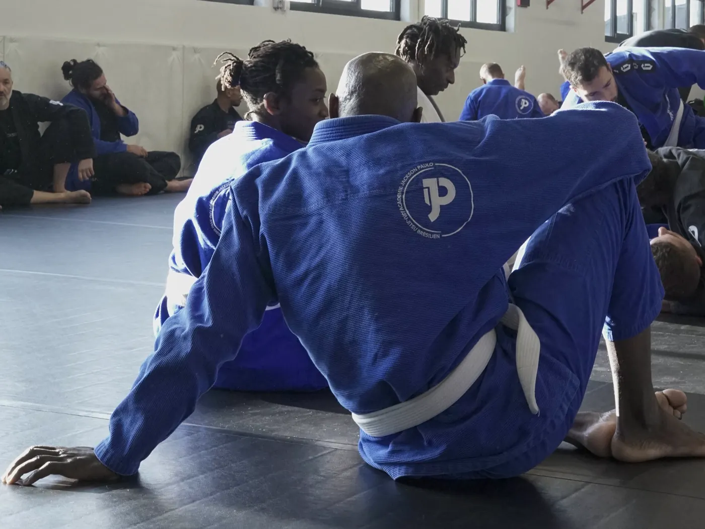 Un élève en kimono bleu et ceinture blanche, vu de dos, assis sur le tapis entre deux exercices